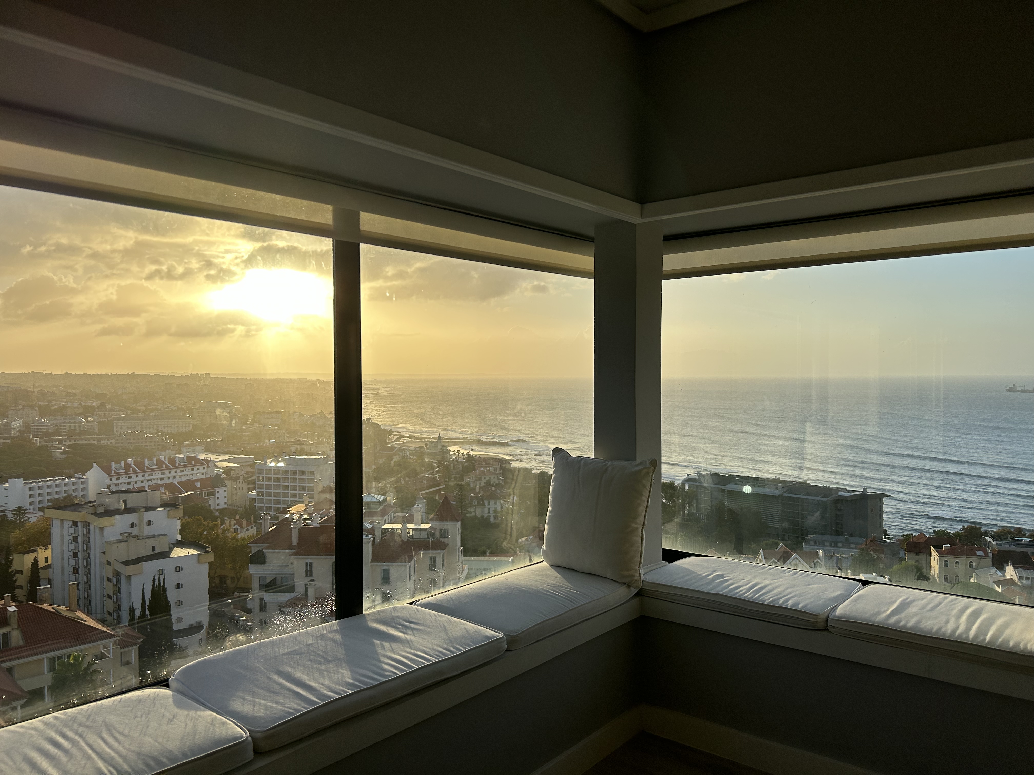 Morning view from a high-rise building's corner window seat, with the sun rising above a coastal cityscape. Sunlight streams through the clouds, casting a soft glow over the sea and illuminating the city. The calm ocean extends to the horizon under a sky graced with the early light of day.