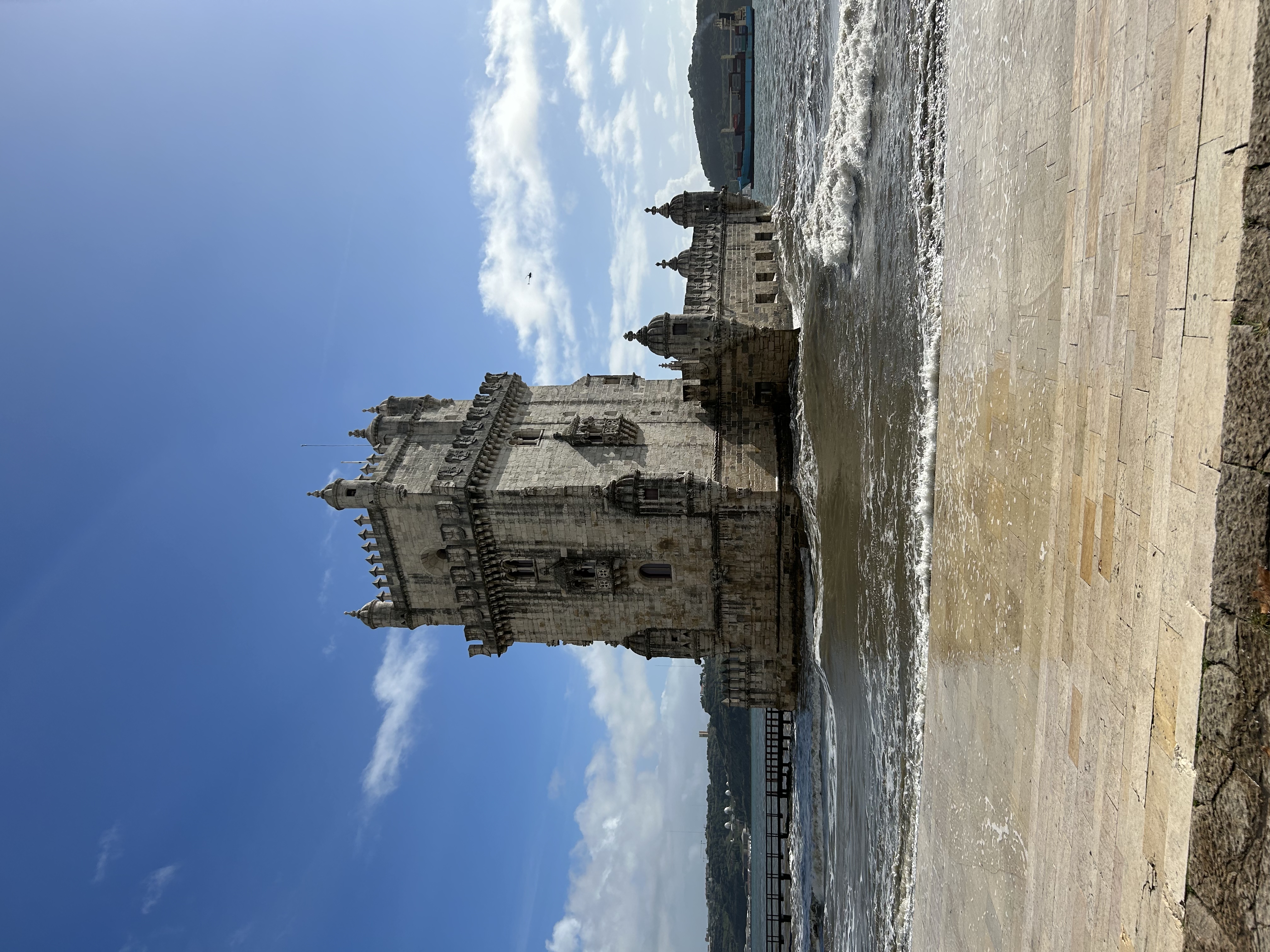 Belem Tower standing majestically under a clear blue sky, its intricate limestone carvings and sturdy battlements highlighted by the bright sunlight. The Tagus River surrounds the tower, with gentle waves lapping against the stone embankment in the foreground.