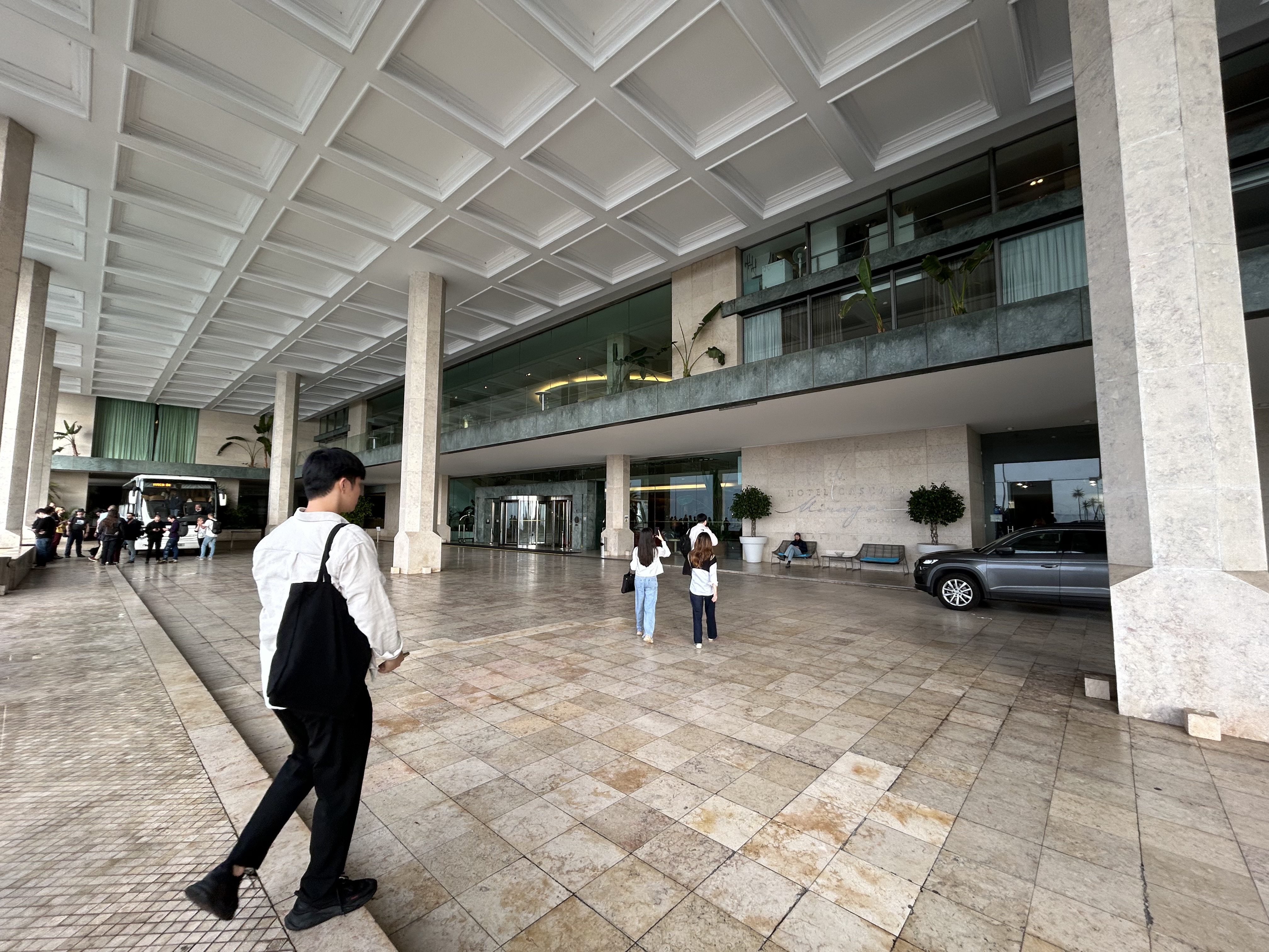 Entrance of a modern conference venue with a high ceiling supported by square columns. People are scattered around the space, some walking and others standing in groups, chatting. A man in a white shirt and black trousers walks across the foreground, while a car is parked by the curb, and the interior of the building is visible through the glass facade in the background.
