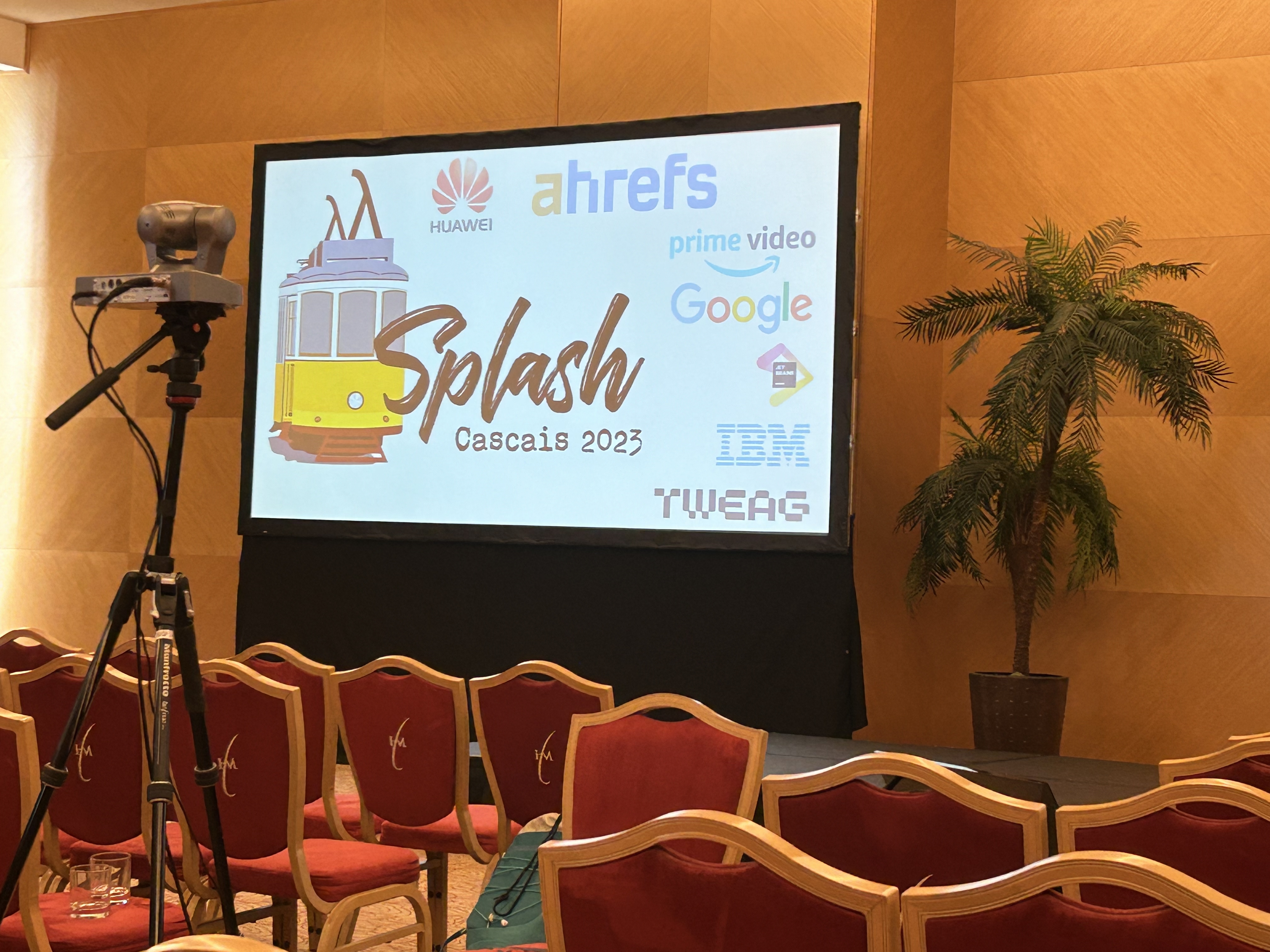 Conference room setup for the Splash event in Cascais 2023, with a large projector screen displaying the event's title and logos of sponsors like Huawei, Ahrefs, Prime Video, Google, and IBM. Red chairs with curved backs are neatly arranged in rows, facing the screen. A camera on a tripod stands to the left, ready to record, and a potted palm tree adds a touch of greenery to the right side of the room.